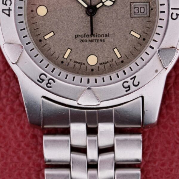 Close-up of a silver Tag Heuer 1500 Series watch with a round face, showing the date at 3 oclock. It reads professional 200 METERS and T SWISS MADE T, with a metal link bracelet against a textured red background.