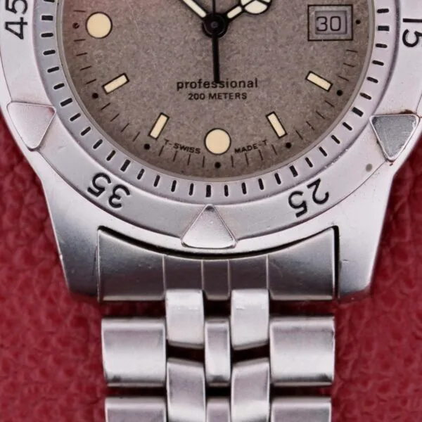 Close-up of a silver Tag Heuer 1500 Series watch with a round face, showing the date at 3 oclock. It reads professional 200 METERS and T SWISS MADE T, with a metal link bracelet against a textured red background.