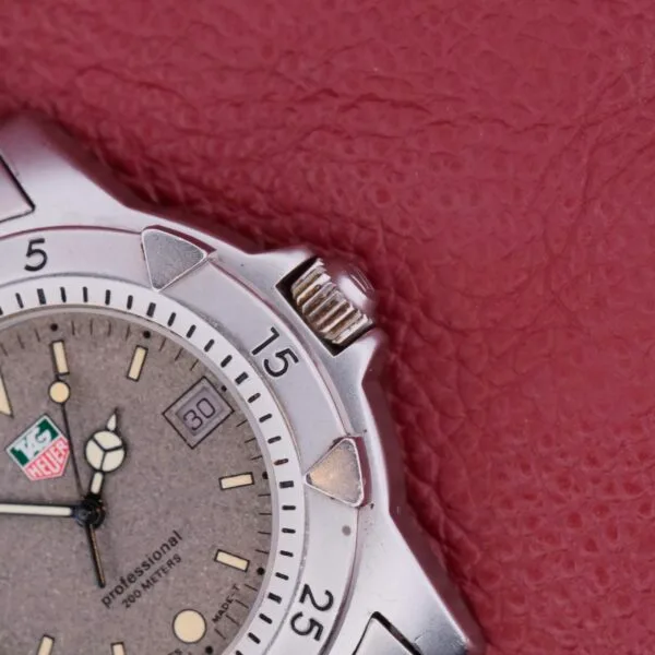 Close-up of a silver Tag Heuer 1500 Series watch on a textured red surface, featuring a round face with time, minute markers, and date display. The bezel has numbers, and the crown is visible on the right side.