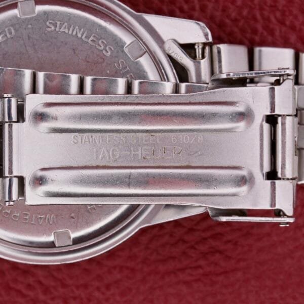 A close-up shows a metallic watch clasp stamp-marked with STAINLESS STEEL 610/9 TAG HEUER against a red textured backdrop, highlighting the intricate detailing and secure locking mechanism of the iconic Tag Heuer 1500 Series.
