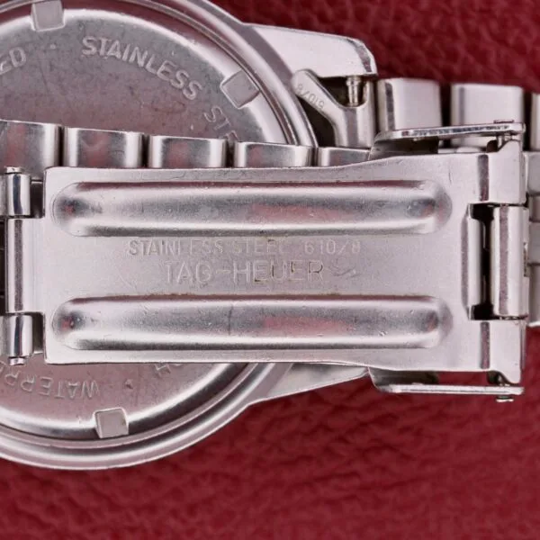 A close-up shows a metallic watch clasp stamp-marked with STAINLESS STEEL 610/9 TAG HEUER against a red textured backdrop, highlighting the intricate detailing and secure locking mechanism of the iconic Tag Heuer 1500 Series.