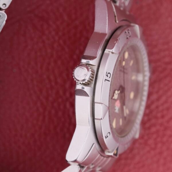 Close-up of a sleek silver Tag Heuer 1500 Series wristwatch on a red background, showcasing its side profile. The round dial displays time markers, a crown, a small date window, and the iconic logo.