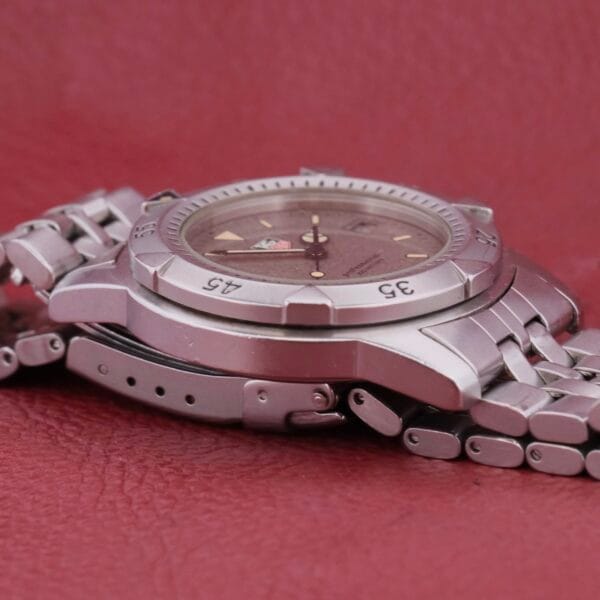 A silver Tag Heuer 1500 Series wristwatch with a metallic band rests on its side against a textured maroon background, featuring a dark face with visible numerals on the beveled edge.