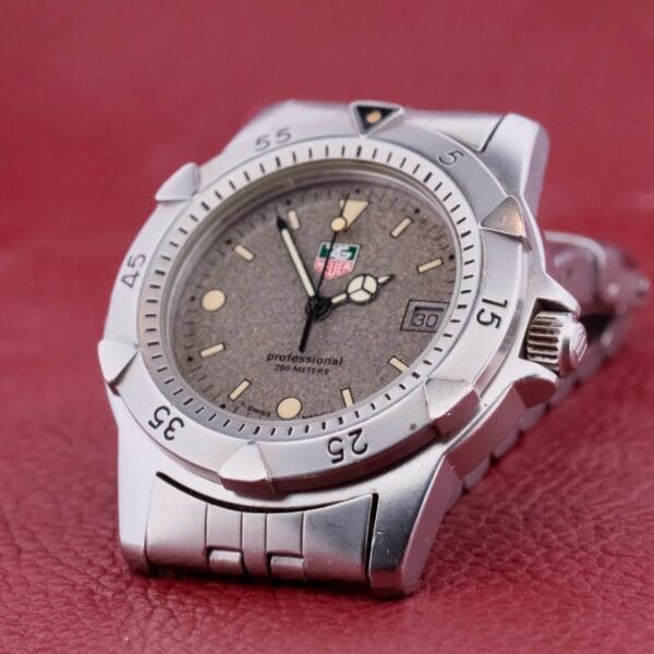 The Tag Heuer 1500 Series features a silver wristwatch with a gray textured dial, date display, and luminescent markers. Its bezel has minute indicators that contrast with the red leather background.
