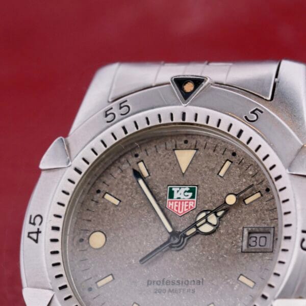 Close-up of a Tag Heuer 1500 Series professional divers watch showcasing a textured silver dial, luminescent hands, date window at 3 oclock, and unidirectional bezel with minute markers. Water resistant up to 200 meters.