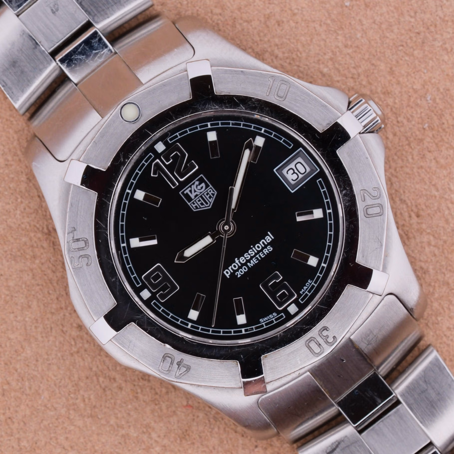 A Tag Heuer Professional Dive wristwatch with a black dial, date display, and metal band rests on a tan surface. TAG Heuer Professional 200m ref WN1110-0 front view, black dial with applied steel markers and 12-6-9 Arabic numerals, date at 3, scalloped steel diver bezel