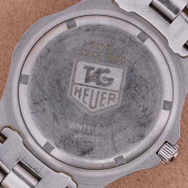 A scratched back of a TAG Heuer Professional Dive watch shows the iconic logo, SWISS SINCE 1860, and model numbers WN1110 and VP8021. It features a sleek metal bracelet.
