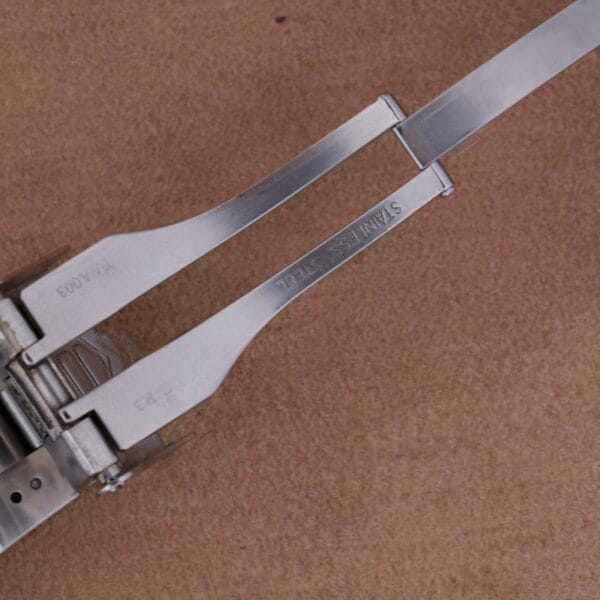 Close-up of a stainless steel hinge on a brown surface, echoing the precision of Tag Heuer Professional Dive watches. The hinge features two parallel arms marked STAINLESS STEEL, highlighting intricate metalwork with visible screws and joints.