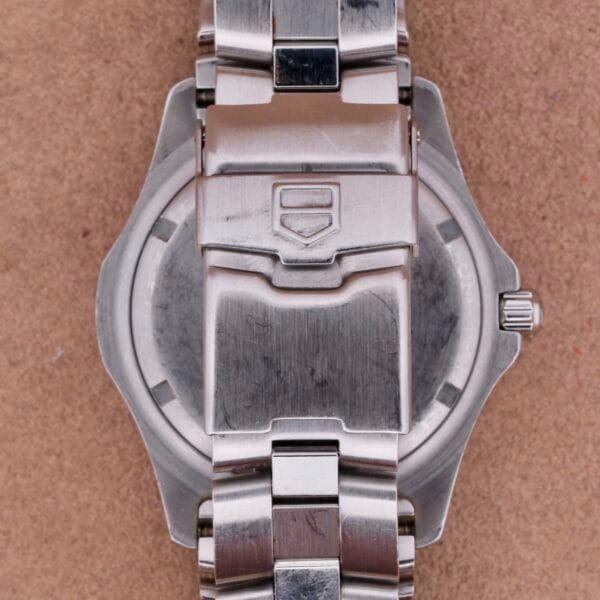 A close-up shows the back of a Tag Heuer Professional Dive watch on a beige surface, with its round stainless steel case and metal bracelet visible.