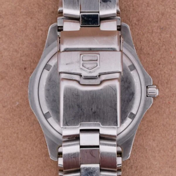 A close-up shows the back of a Tag Heuer Professional Dive watch on a beige surface, with its round stainless steel case and metal bracelet visible.