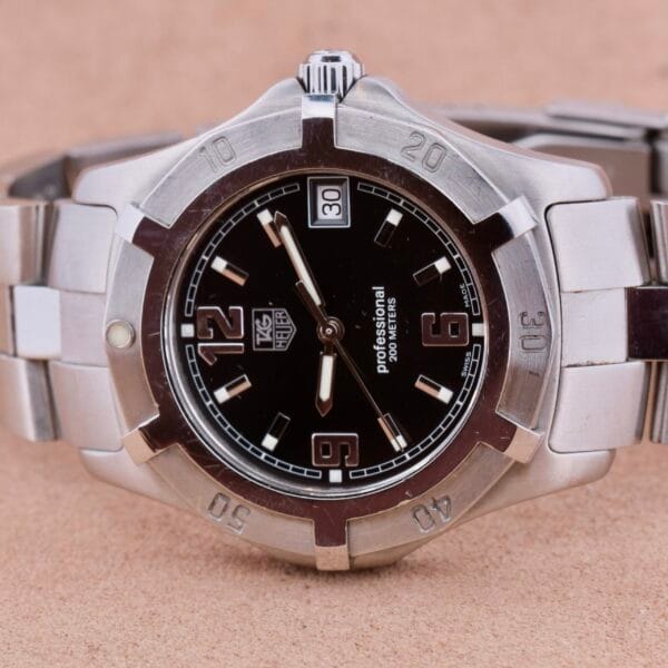 The iconic TAG Heuer Professional Dive watch is showcased with its sleek stainless steel band, a black face with large silver numbers, a date window at 3 oclock, and an engraved unidirectional bezel, all stylishly displayed on a beige surface.