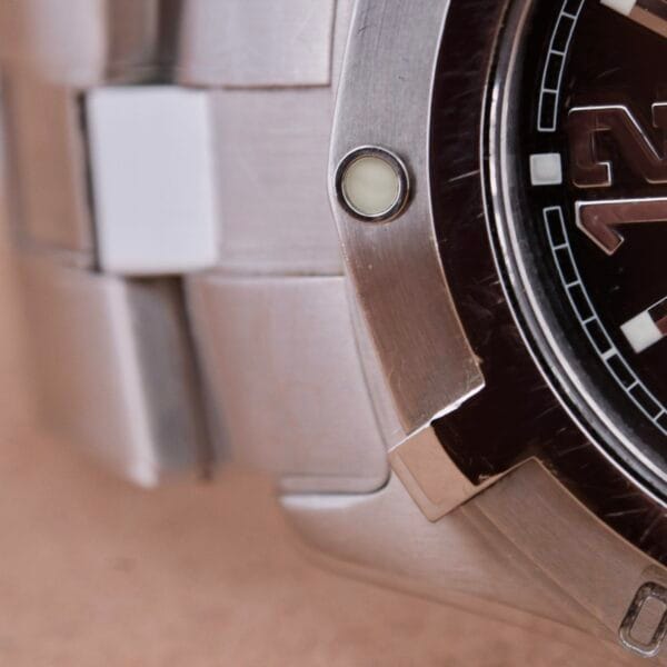Close-up of a Tag Heuer Professional Dive watch. It features a dark face with white markers and a partially visible frame, two screws on the edge, and a subtly shimmering textured metallic band in the background.
