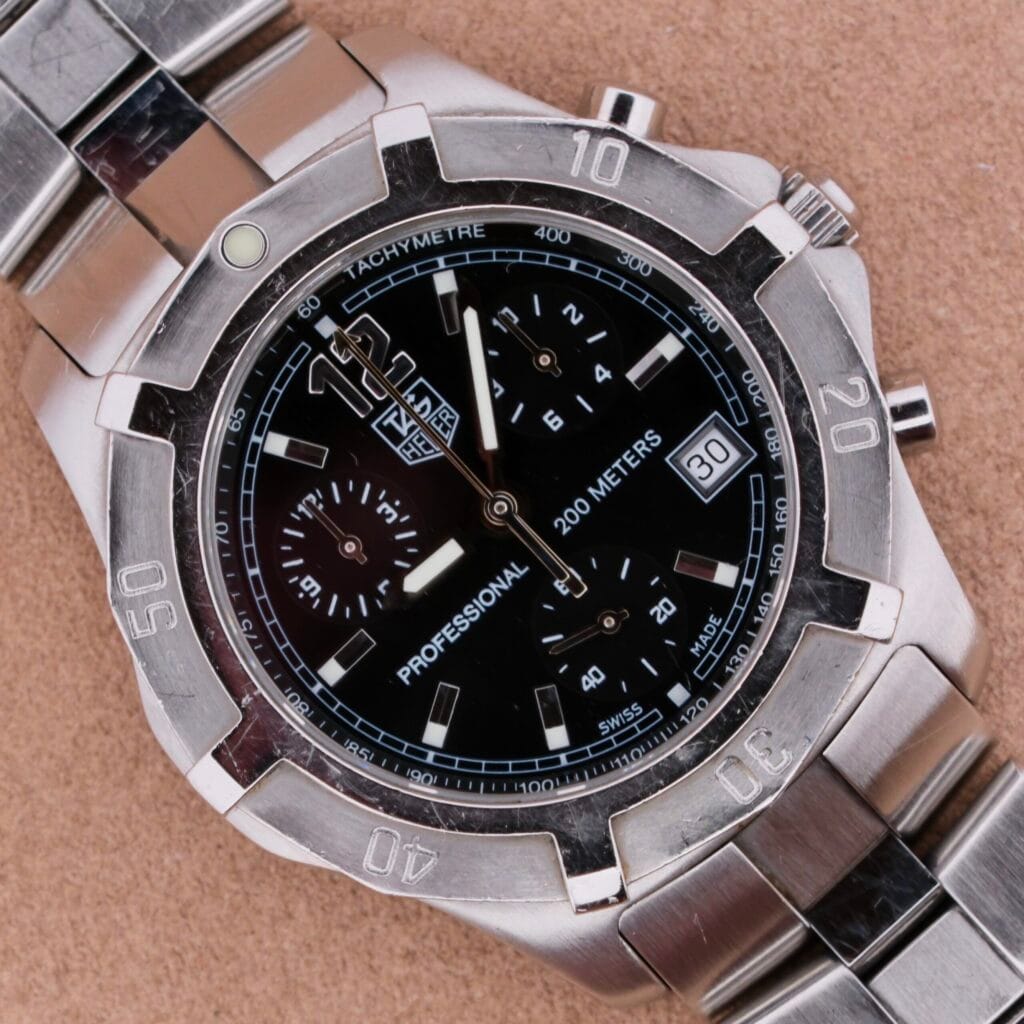 Silver Tag Heuer 2000 Professional Chronograph wristwatch featuring a black dial, date display, and tachymeter bezel resting on a tan surface.