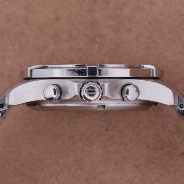 Side view of a Tag Heuer 2000 Professional Chronograph wristwatch with a round case, two push buttons, and a central crown. It rests on a brown textured background, highlighting its sleek metallic design.