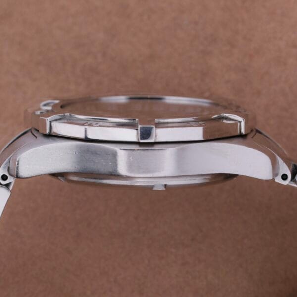 Close-up side view of a silver wristwatch similar to a Tag Heuer 2000 Professional Chronograph, with a polished metal case and lugs, set against a plain brown background. The watch face is hidden from view.