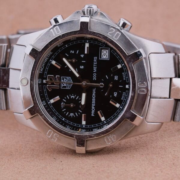 The Tag Heuer 2000 Professional Chronograph features a silver wristwatch with a black dial, tachymeter scale, three subdials, date window, and Professional 200 Meters on the case. It includes a strong metallic strap and is elegantly displayed on a brown background.