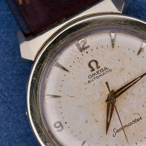 A close-up of the Omega Seamaster Bull Horn Lugs watch, featuring a vintage white dial with patina. It showcases gold hour and minute hands, silver hour markers, and Omega Automatic Seamaster text. The dark leather band enhances its classic charm.