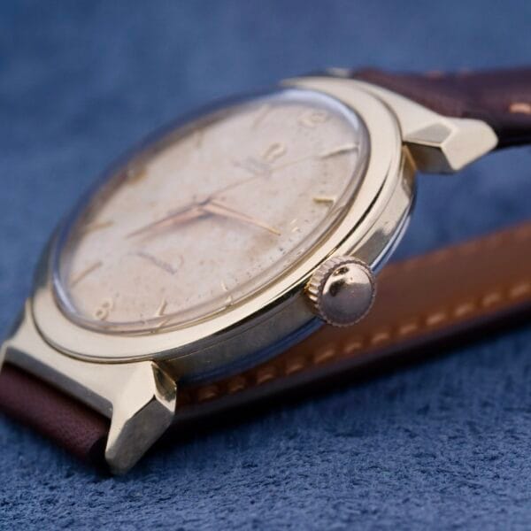 Close-up of a vintage Omega Semaster Bull Horn Lugs wristwatch with a textured off-white dial and brown leather strap, featuring slender hands, small hour markers, and highlighting the side profile with bull horn lugs and crown.