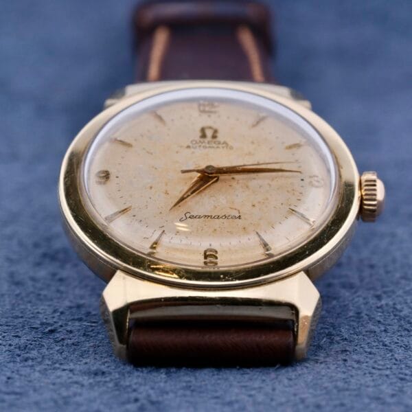 The Omega Seamaster Bull Horn Lugs is a vintage wristwatch featuring a textured cream dial with hour markers and hands, complemented by distinctive bull horn lugs, a brown leather strap, and displayed on a blue textured surface.