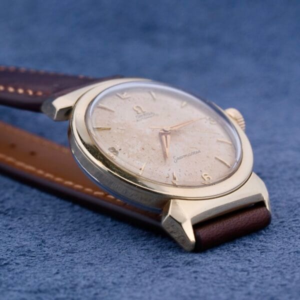 A close-up of the Omega Semaster Bull Horn Lugs wristwatch reveals a vintage gold design with leather strap on a blue textured background. The slightly worn watch face features visible hands and hour markers, with Omega and Seamaster subtly inscribed, enhancing its timeless allure.