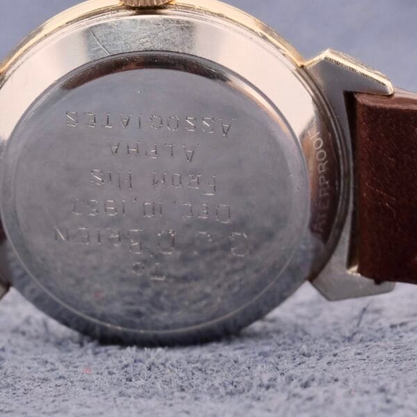 Close-up of the back of an Omega Semaster Bull Horn Lugs wristwatch on a textured gray surface. The bull horn lugs enhance its metal case and brown leather strap. The text on the back, partially visible, includes a date, suggesting personalized engraving.