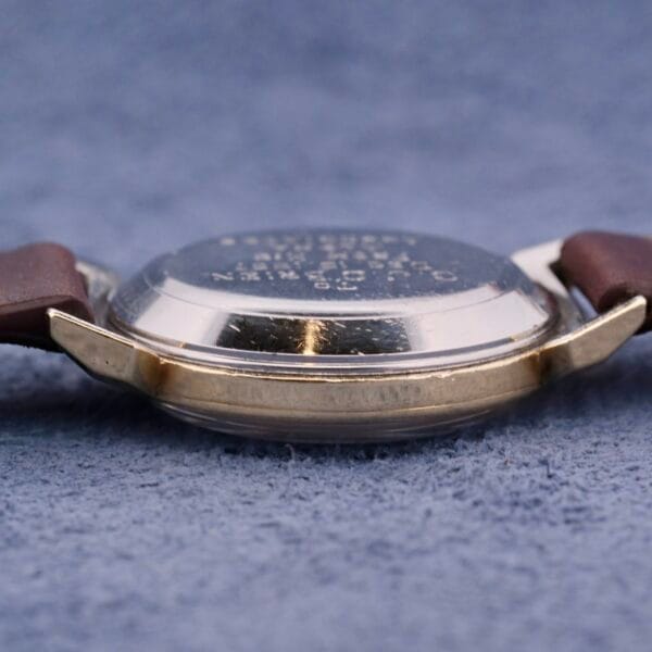 A silver Omega Seamaster Bull Horn Lugs wristwatch with a brown leather strap rests on soft blue fabric. Positioned on its side, it reveals the engraved text on the back case and its slightly reflective surface.
