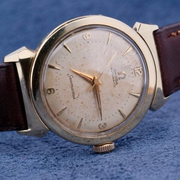 The Omega Seamaster Bull Horn Lugs is a vintage watch with a gold case, white dial, visible patina, and a brown leather strap against a blue textured backdrop.