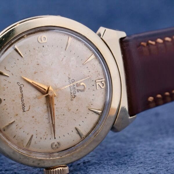 A close-up of the Omega Seamaster Bull Horn Lugs watch highlights its iconic lugs, worn cream-colored dial, gold hour markers, gold case, and brown leather strap. The timepiece is elegantly set against a textured blue surface.