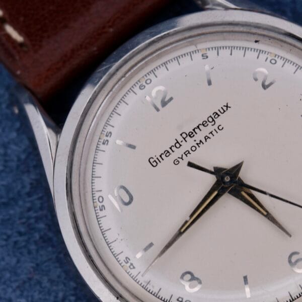 Close-up of a Girard Perregaux Gyromatic wristwatch featuring a silver case, white dial with black numerals and hands, set against a blue background. The elegant design is completed with a brown leather strap.