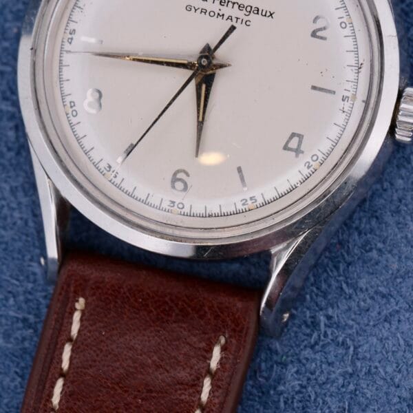 A close-up of a Girard Perregaux Gyromatic watch with a silver case, white dial, brown leather strap, black hands and numerals, and a date window on the dial. It rests on a textured blue surface.