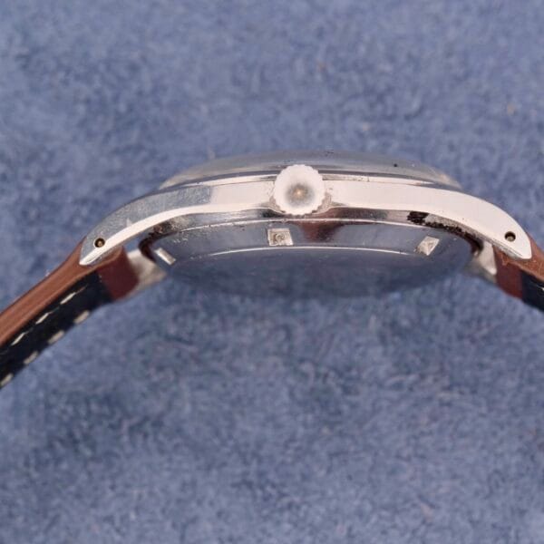 Side view of the Girard Perregaux Gyromatic 6169 wristwatch shows a silver round case and black leather strap with white stitching, set against a textured blue fabric background.