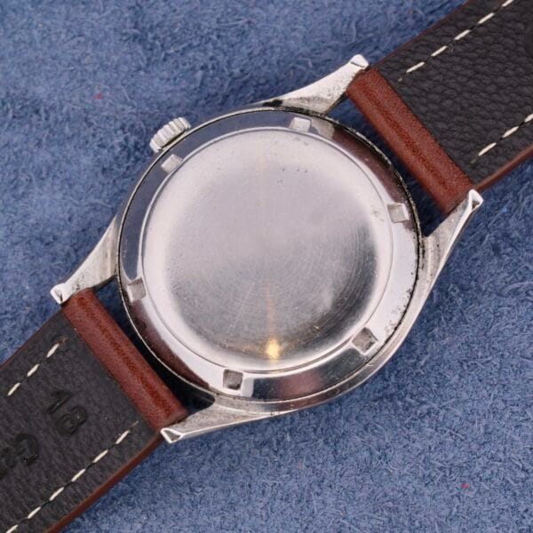 A close-up of a Girard Perregaux Gyromatic wristwatch model 6169 features a silver case and brown leather strap, set against a textured blue background. The watch back is plain with a smooth, reflective finish.