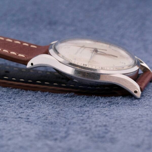 Close-up of a Girard Perregaux Gyromatic wristwatch featuring a round silver case and brown leather strap with white stitching, resting on soft blue fabric. The watch face is white with subtle details.