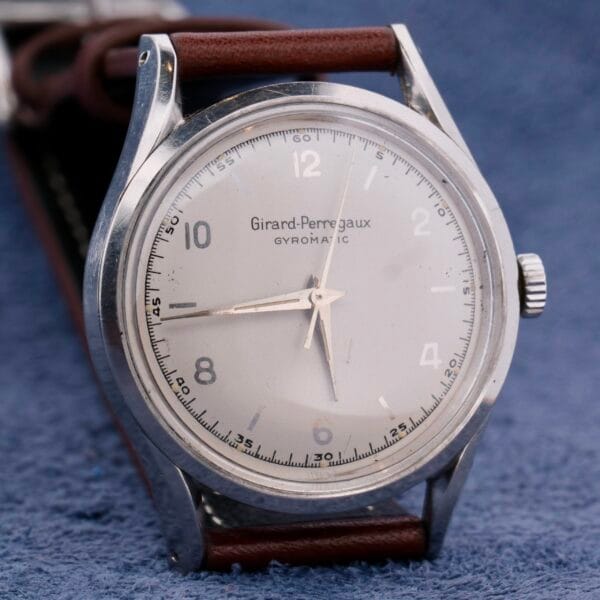 The Girard Perregaux Gyromatic wristwatch features a silver dial with silver hour markers and hands, a minimalistic design, and a brown leather strap, elegantly set against a textured blue background.