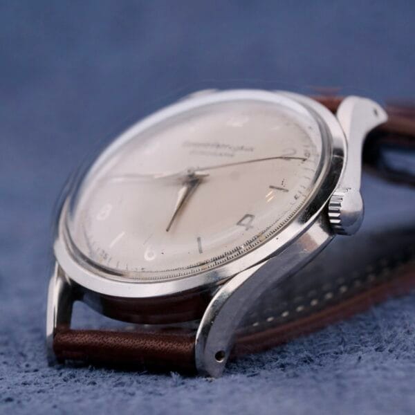 Close-up of a vintage Girard Perregaux Gyromatic wristwatch with a cream dial, silver case, and brown leather strap. The watch has numbers and slender hands, elegantly placed on a soft blue fabric background.