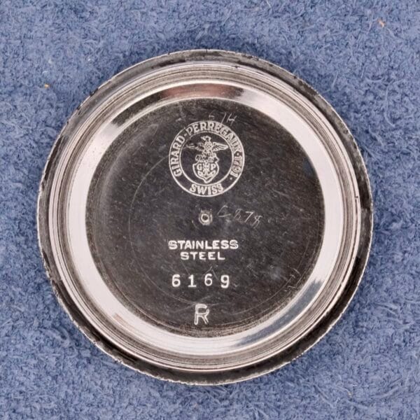 The image reveals a Girard Perregaux Gyromatic watch case back on a blue textured background, highlighting the Swiss emblem. It features Stainless Steel, the number 6169, and showcases its precision with intricate markings and engravings.