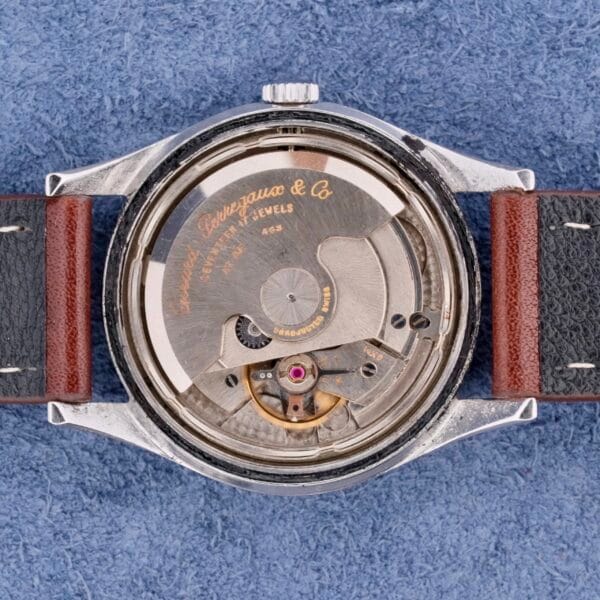 The image reveals the back view of a Girard Perregaux Gyromatic, showcasing its exposed mechanical components. The engraved movement is the Gyromatic 6169. The watch rests on a blue textured surface and is fitted with a two-tone leather strap.