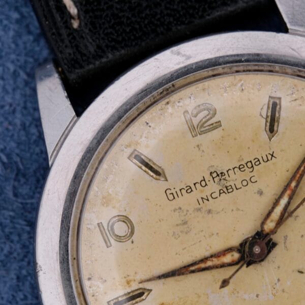 Close-up of a vintage Girard Perregaux Incabloc Manual Wind watch face, highlighting its worn cream dial with Arabic numerals at 12, 3, and 9. The watch features a metal case and black leather strap against a textured blue background.