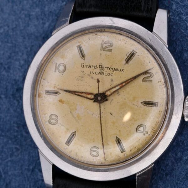 The vintage Girard Perregaux Incabloc Manual Wind watch has a worn cream-colored dial, silver hands, Arabic numeral hour markers, a metal case, and a black leather strap, elegantly displayed on a textured blue surface.