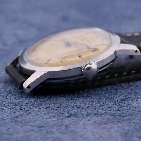 Close-up of a vintage Girard-Perregaux Incabloc Manual Wind wristwatch on a textured blue surface. It has a worn round metal case, domed crystal, black leather strap with white stitching, and a prominently visible crown on the side.