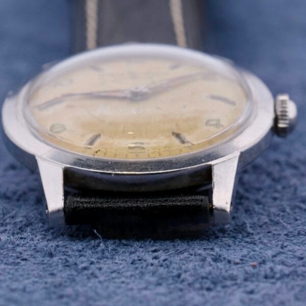 Close-up of a vintage Girard Perregaux Incabloc Manual Wind watch, featuring a worn beige dial and black leather strap on a blue textured surface. The metal case shows slight tarnishing, reflecting its age.