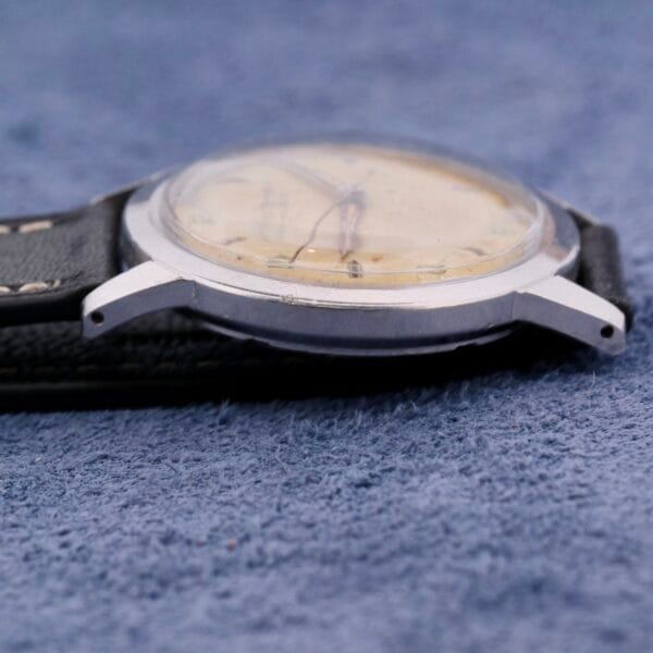Close-up of a vintage Girard Perregaux Incabloc Manual Wind watch with a worn, yellowed face and black leather strap on a textured blue surface. The metal casing shows scratches and aging.