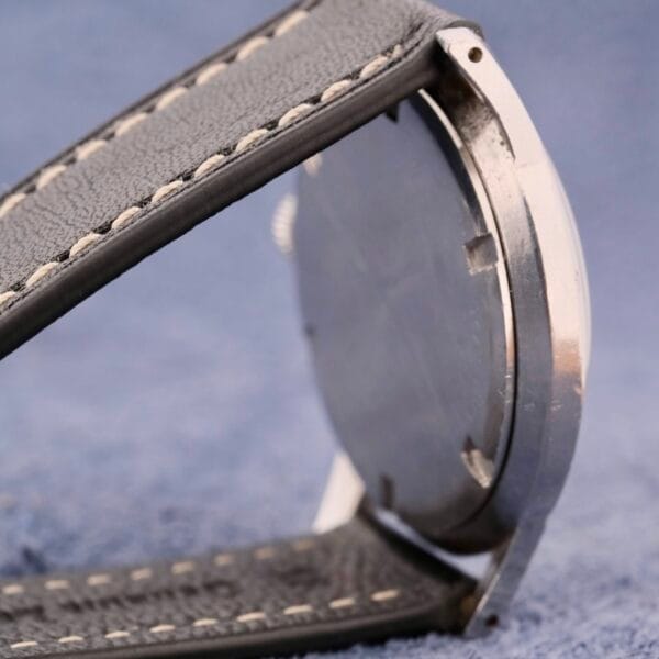 A close-up side view captures the elegance of a Girard Perregaux Incabloc Manual Wind watch on a blue surface. The focus is on its grey leather strap with white stitching and metallic case, highlighting refined craftsmanship while keeping the watch face hidden for an air of mystery.
