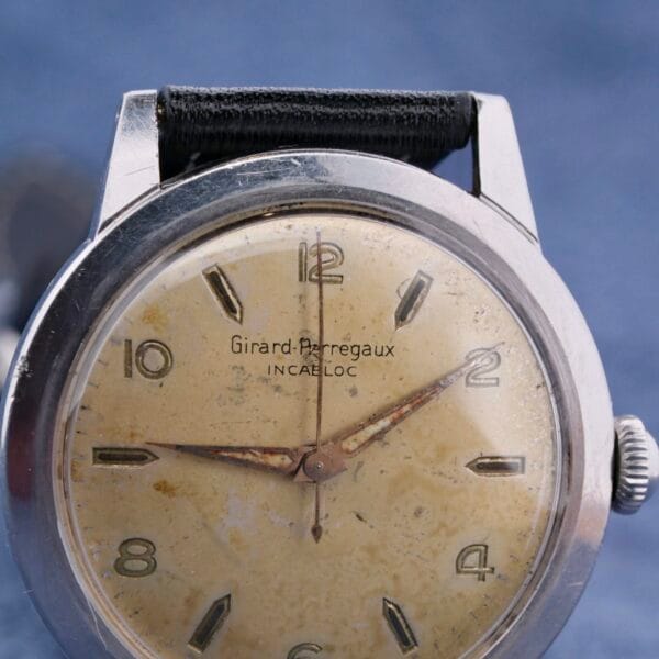 A vintage Girard Perregaux Incabloc Manual Wind watch with a worn cream dial, Arabic numerals, and aged patina hands. It features a silver case and is complemented by a black textile strap.