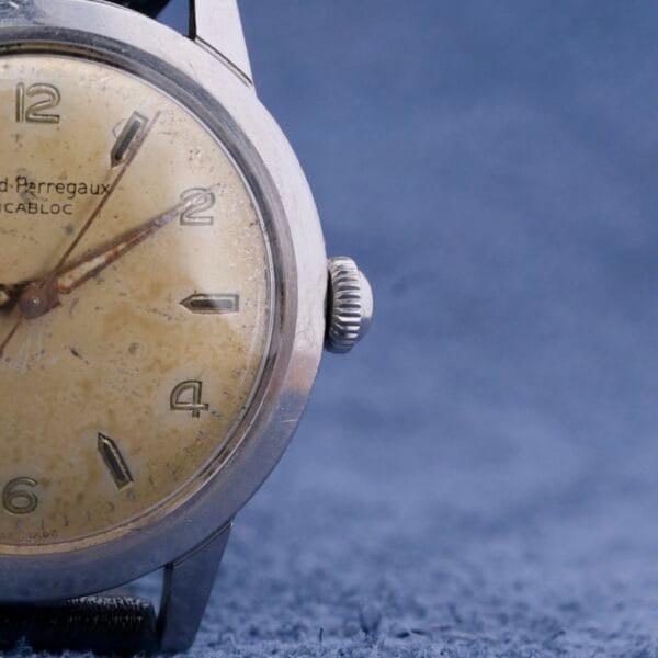 Close-up of a Girard Perregaux Incabloc Manual Wind watch with an aged beige dial, silver case, and Arabic numerals indicating 4:38. The soft, textured blue background enhances the timepieces classic charm.