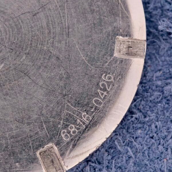 Close-up of a scratched metal surface with engraved numbers 6816 0426, resembling the detailed craftsmanship of a Girard Perregaux Incabloc Manual Wind watch, adorned with four silver tabs on a textured blue fabric background.