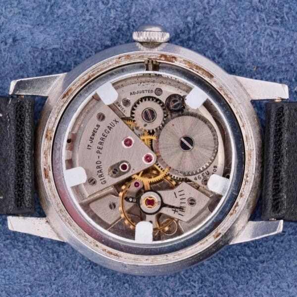 The Girard-Perregaux Incabloc Manual Wind watch features a vintage mechanical design with an open-back case revealing intricate gears, a silver casing labeled Girard-Perregaux, visible jewels, and a black leather strap set against a blue fabric background.
