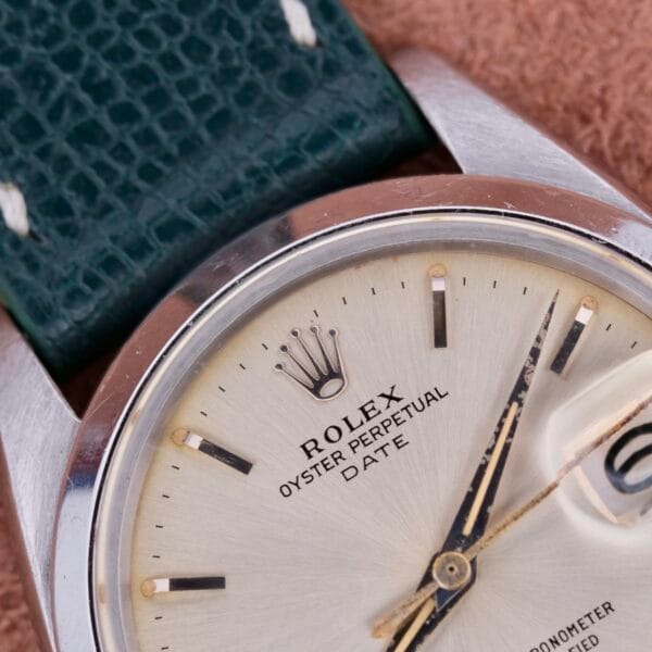 The Rolex Oyster Perpetual Date 1500 Silver Dial showcases its elegant silver dial with silver hour markers and hands. Complemented by a green leather strap, it features a date window with cyclops lens below the iconic Rolex crown logo.