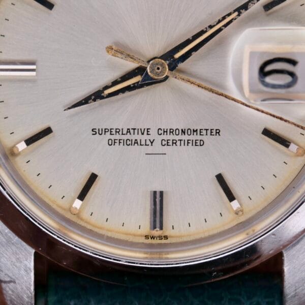 Close-up of a Rolex Oyster Perpetual Date 1500 Silver Dial watch face with silver hands and black hour markers showing the number 6 in the date window. The text reads SUPERLATIVE CHRONOMETER OFFICIALLY CERTIFIED. It features a polished metal casing.