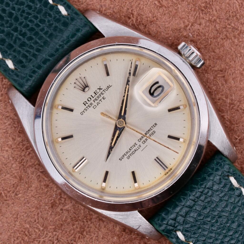 A close-up of a Rolex Oyster Perpetual Date 1500 Silver Dial watch shows its cream dial, magnified date window at 6, and elegant silver case. The green leather strap contrasts luxuriously with the brown textured background.